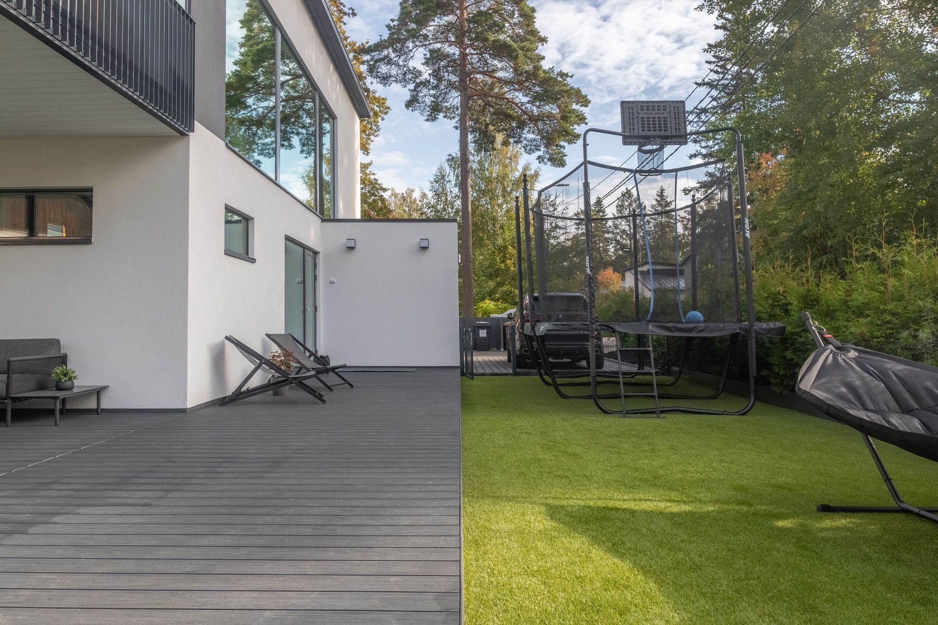 Moderni takapihan patio harmaalla terassilla, vihreällä nurmikolla, trampoliinilla ja lepotuoleilla valkoisen talon vieressä