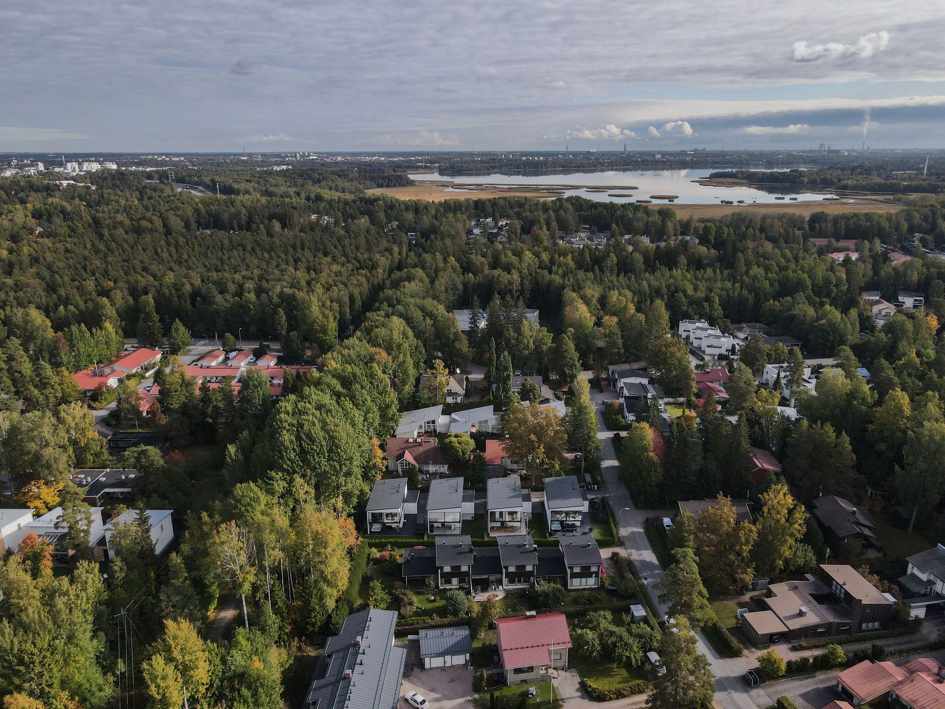 Ilmakuva metsäisestä esikaupunkialueesta, jossa on hajallaan olevia taloja järven lähellä pilvisen taivaan alla