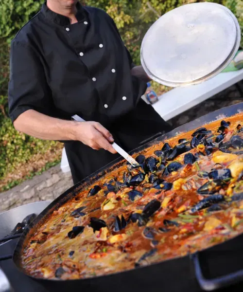 Un chef prépare une paella aux fruits de mer dans une grande poêle à l'extérieur. Il porte un uniforme noir ; on aperçoit des moules et des tomates.