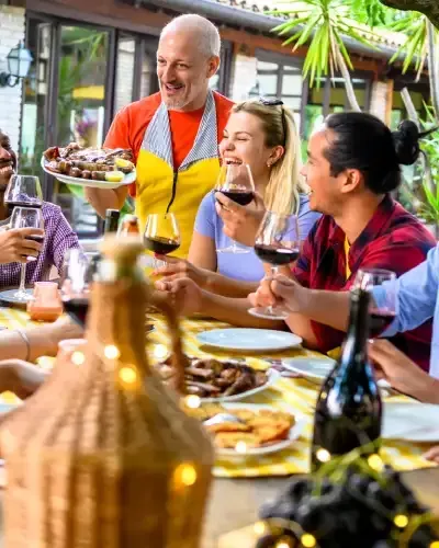 Un groupe de personnes attablées en terrasse ; un homme sert le repas, d’autres rient et boivent du vin.