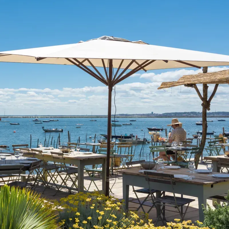 Un restaurant en plein air en bord de mer, des tables dressées sous des parasols, une eau et un ciel bleus.