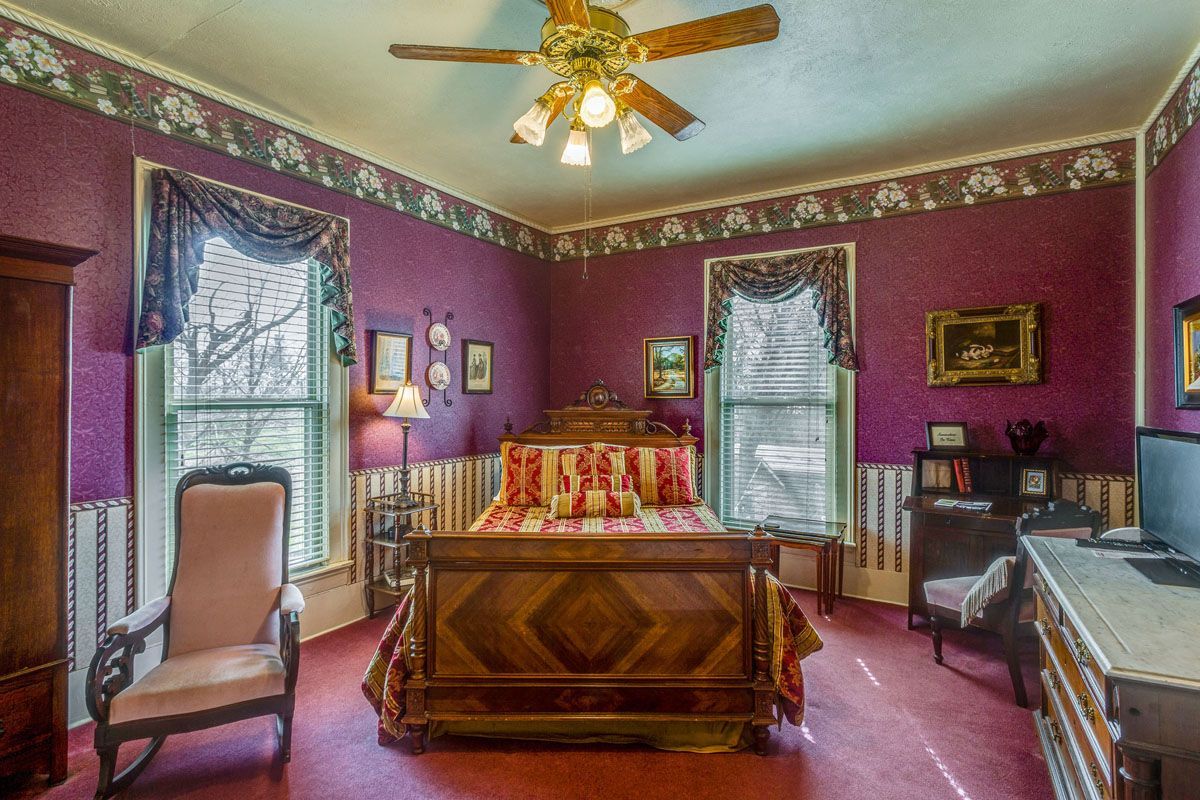 A bedroom with purple walls , a bed , a chair , and a ceiling fan.