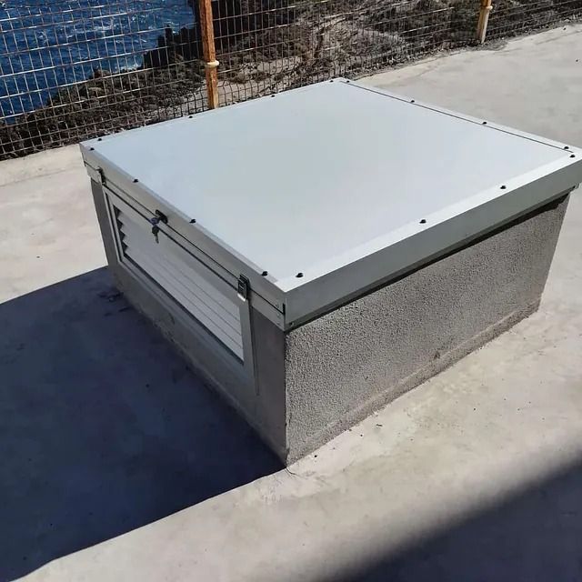 Una caja gris con una tapa de metal está en una acera al lado de una valla.