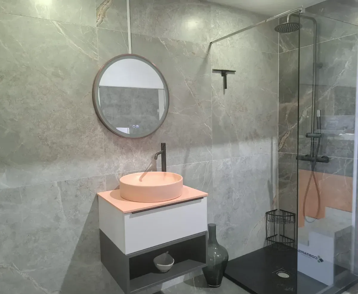 Baño moderno con paredes de azulejos grises, espejo redondo, lavabo color melocotón y ducha de cristal.