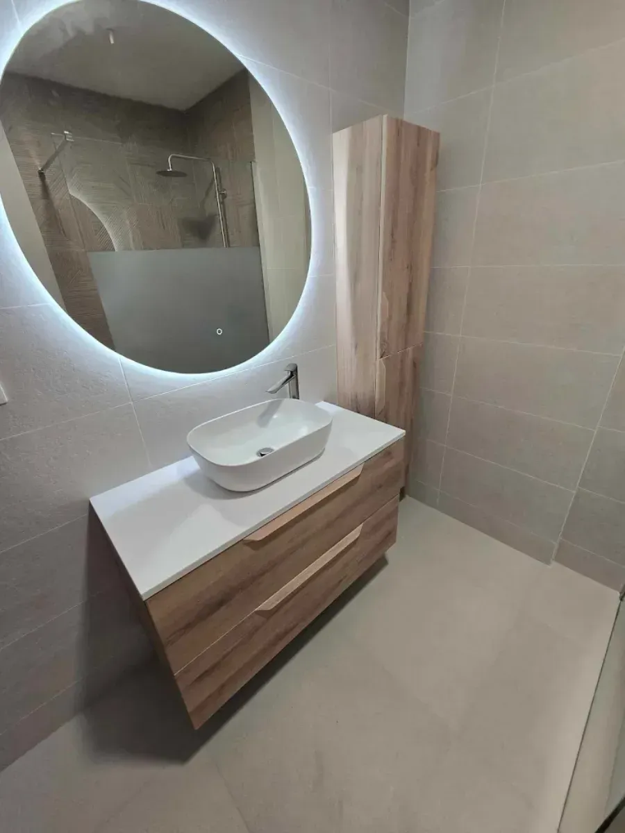 Baño moderno con espejo redondo, tocador de madera y mueble alto.