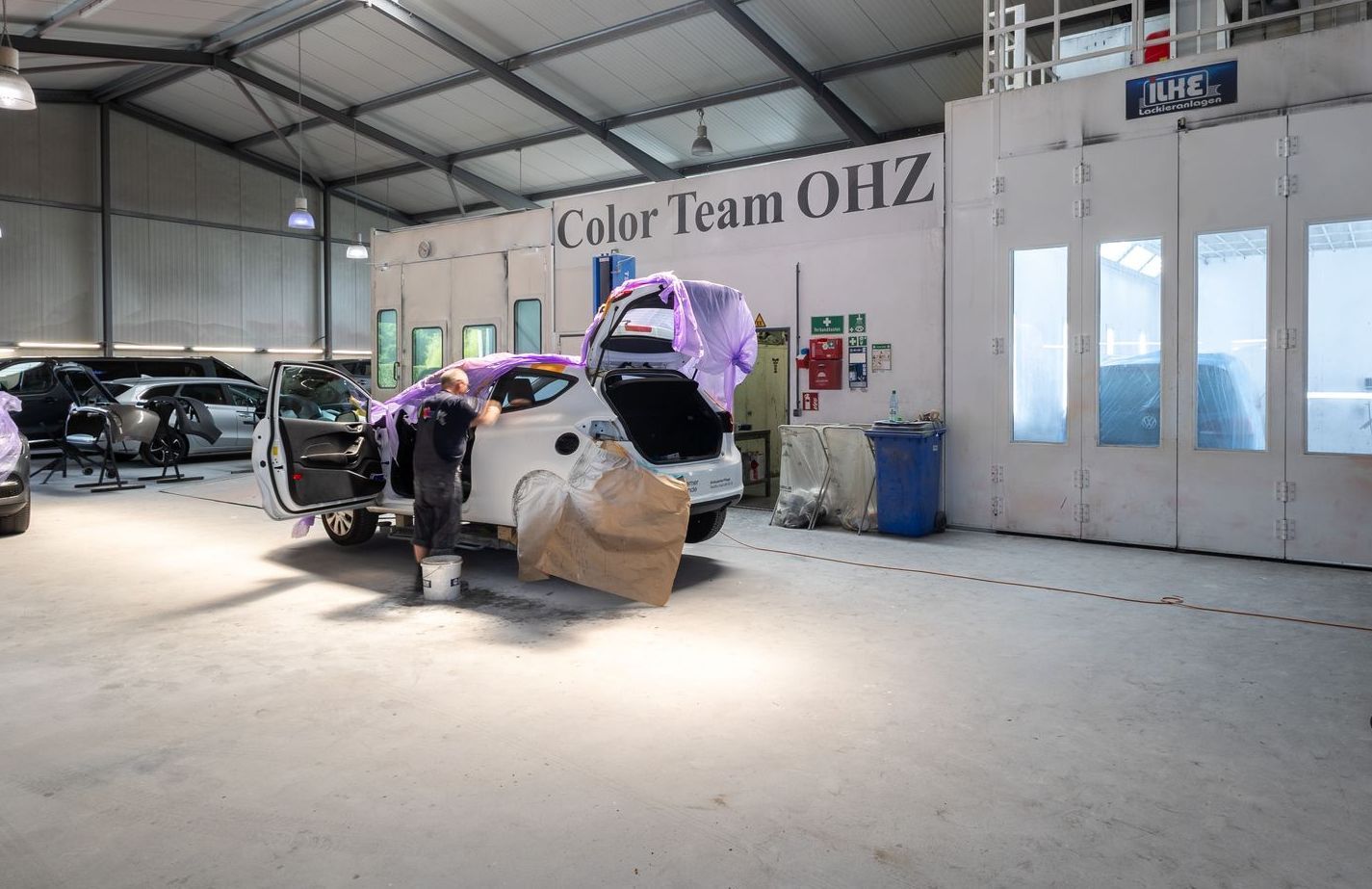 In einer Garage wird ein Auto lackiert, an dem ein Schild mit der Aufschrift „Color Team OHZ“ hängt.