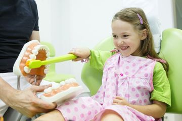 Una niña está sentada en una silla dental cepillándose los dientes con un cepillo de dientes de juguete.
