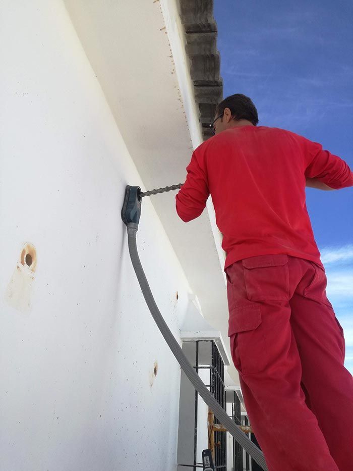 Un trabajador con uniforme rojo pinta una pared blanca con un rodillo en una escalera.