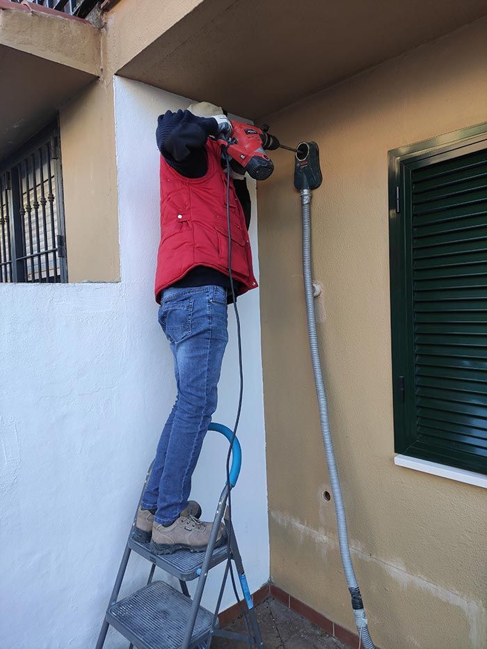 Un trabajador con chaqueta roja está subido a una escalera y utiliza una herramienta eléctrica en una pared exterior.