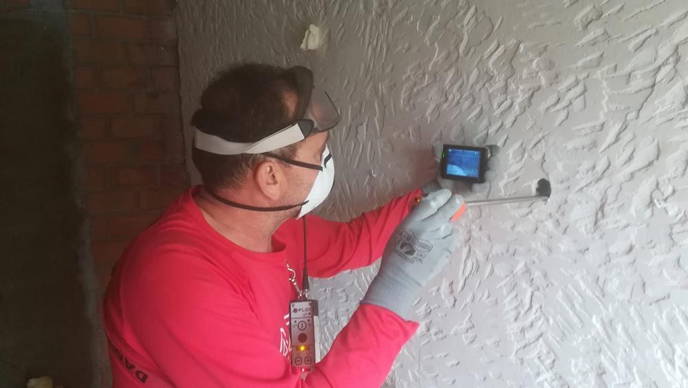 Técnico con camisa roja utilizando un dispositivo portátil para inspeccionar una pared texturizada en el interior.