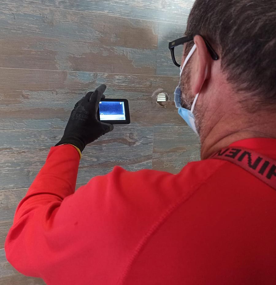 Un trabajador con camisa roja y guantes inspecciona una pared con un dispositivo portátil.