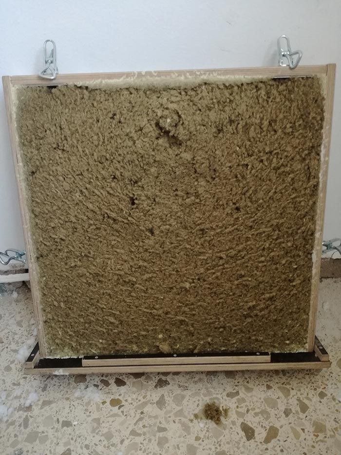 Gran panel cuadrado de color beige con textura, apoyado contra una pared en el suelo, con ganchos en la parte superior.