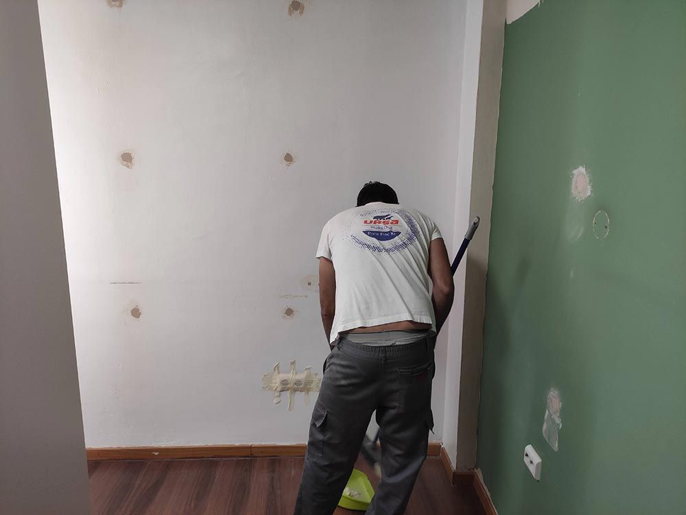 Una persona está pintando una habitación, de pie junto a una pared verde, con una bandeja de pintura y herramientas cerca.
