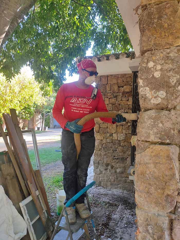 Obrero con camisa roja cincelando un muro de piedra junto a un árbol en una zona de construcción al aire libre.