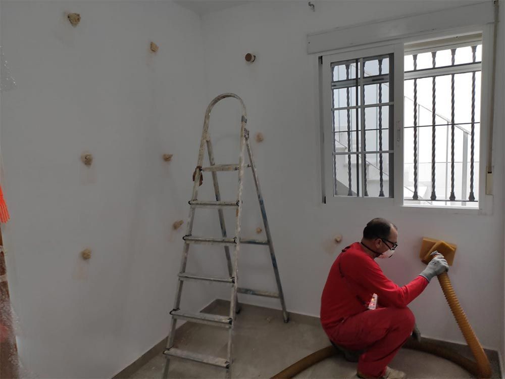 Un trabajador con mono rojo pinta una habitación con una escalera y una ventana enrejada cerca.