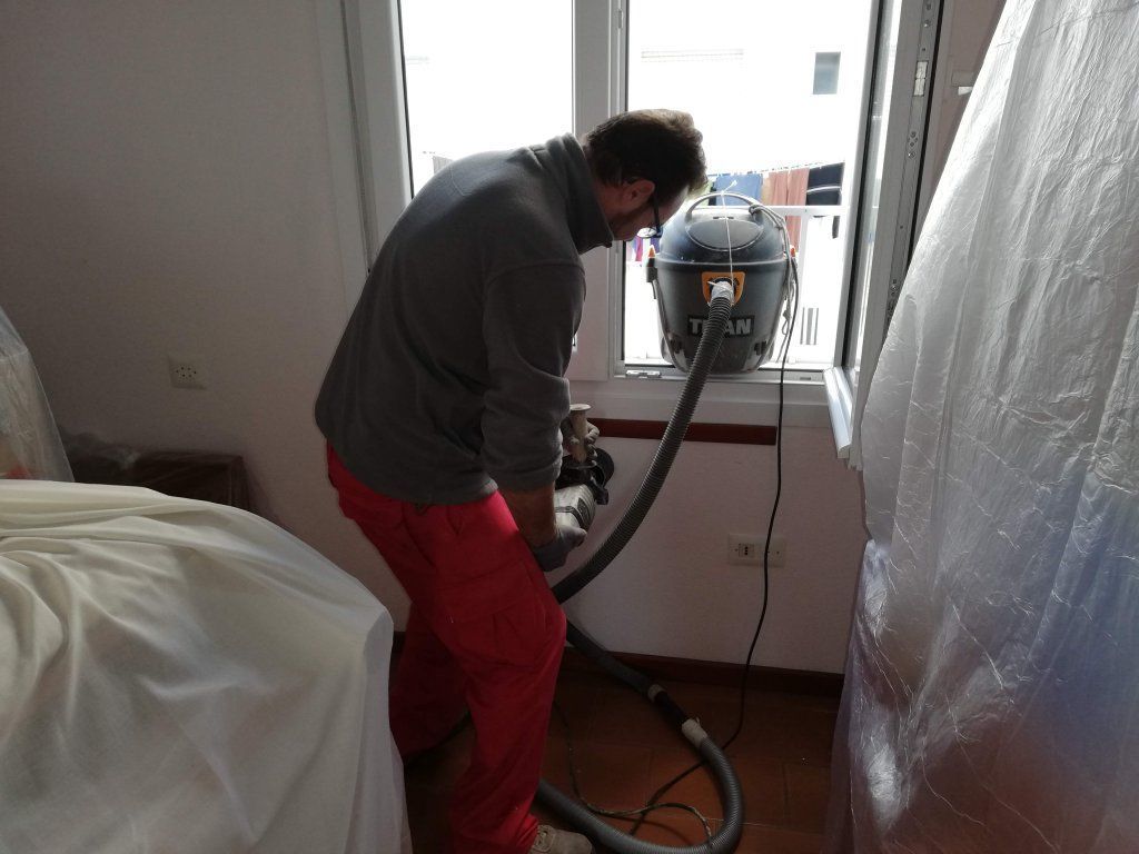 Hombre con pantalones rojos y camisa gris usando una aspiradora cerca de una ventana, con una lámina de plástico cerca.