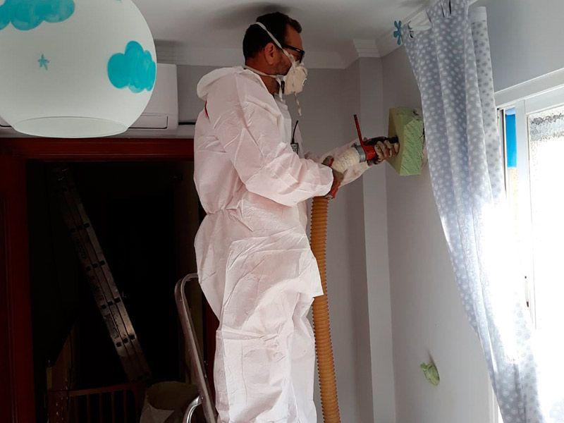 Hombre con traje protector aislando una pared blanca con una manguera, interior de una habitación.