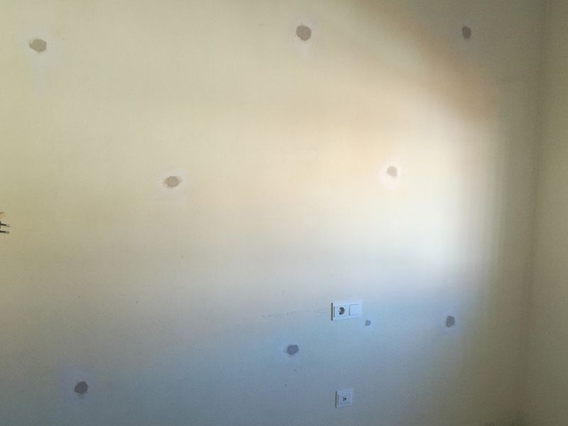 Pared beige con numerosos agujeros tapados de objetos retirados. Tomas de corriente visibles.