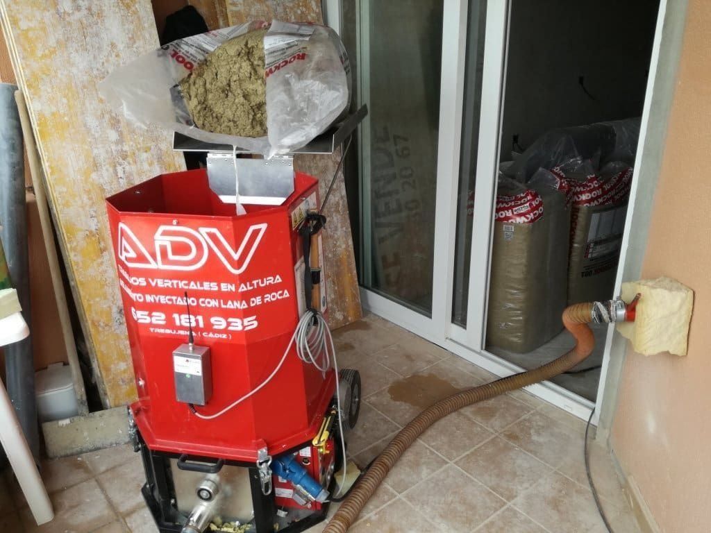 Una máquina roja está inyectando aislamiento en una pared. Hay sacos de aislamiento cerca.