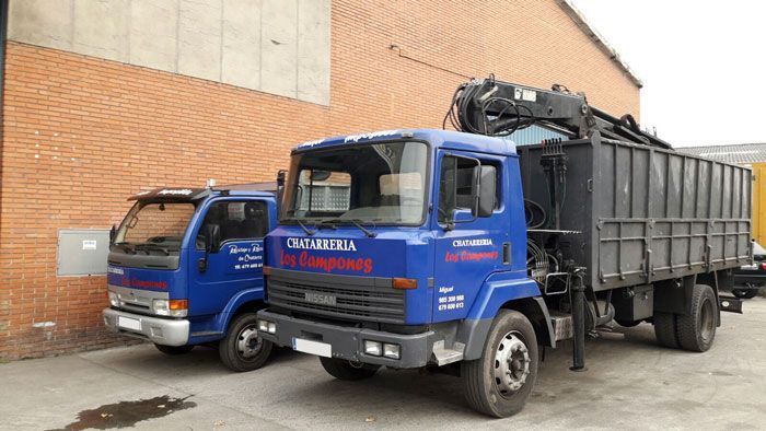 Dos camiones azules estacionados junto a un edificio de ladrillos. El camión más grande lleva una grúa y un contenedor de carga.