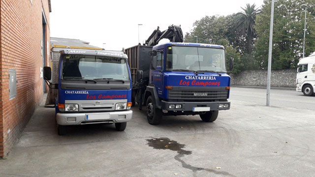 Dos camiones comerciales azules estacionados cerca de un edificio de ladrillo. Uno tiene un elevador de contenedores.