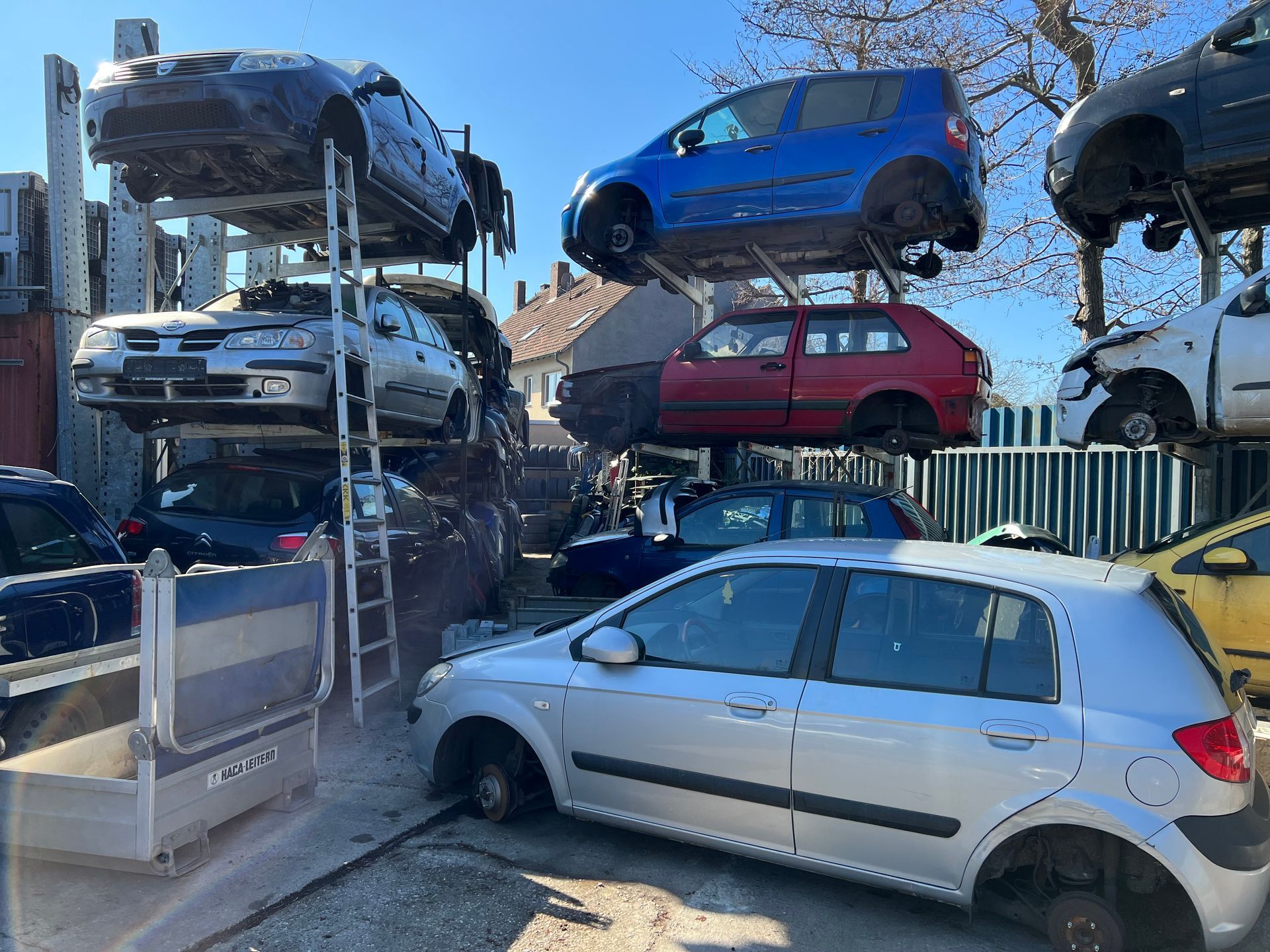 Auf einem Schrottplatz sind gestapelte Autos zu sehen, im Vordergrund ein silberner Schrägheckwagen und auf den oberen Gestellen blaue Autos.