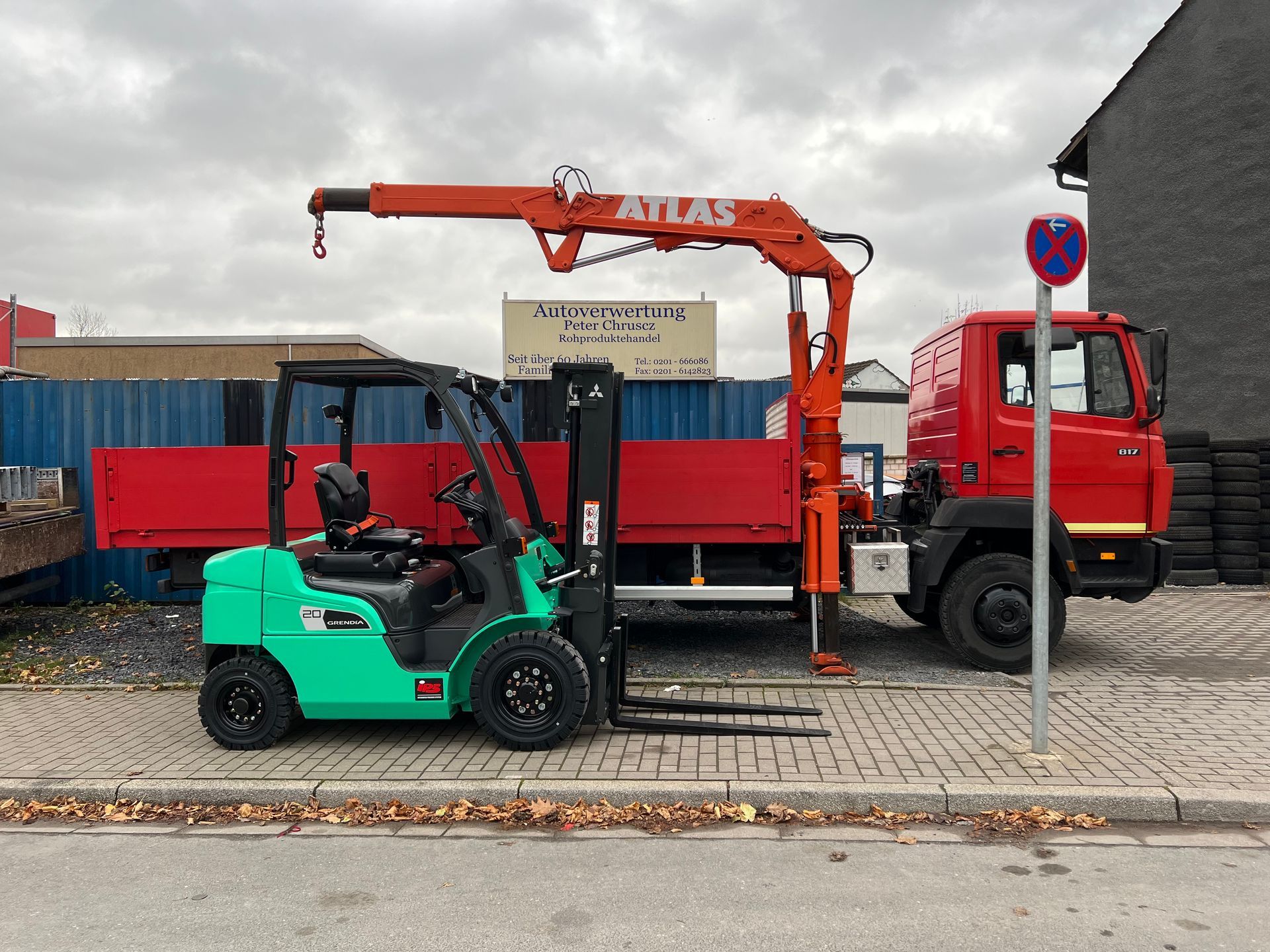 Ein grüner Gabelstapler und ein roter Kranwagen parken neben Schiffscontainern auf einer bewölkten Straße.