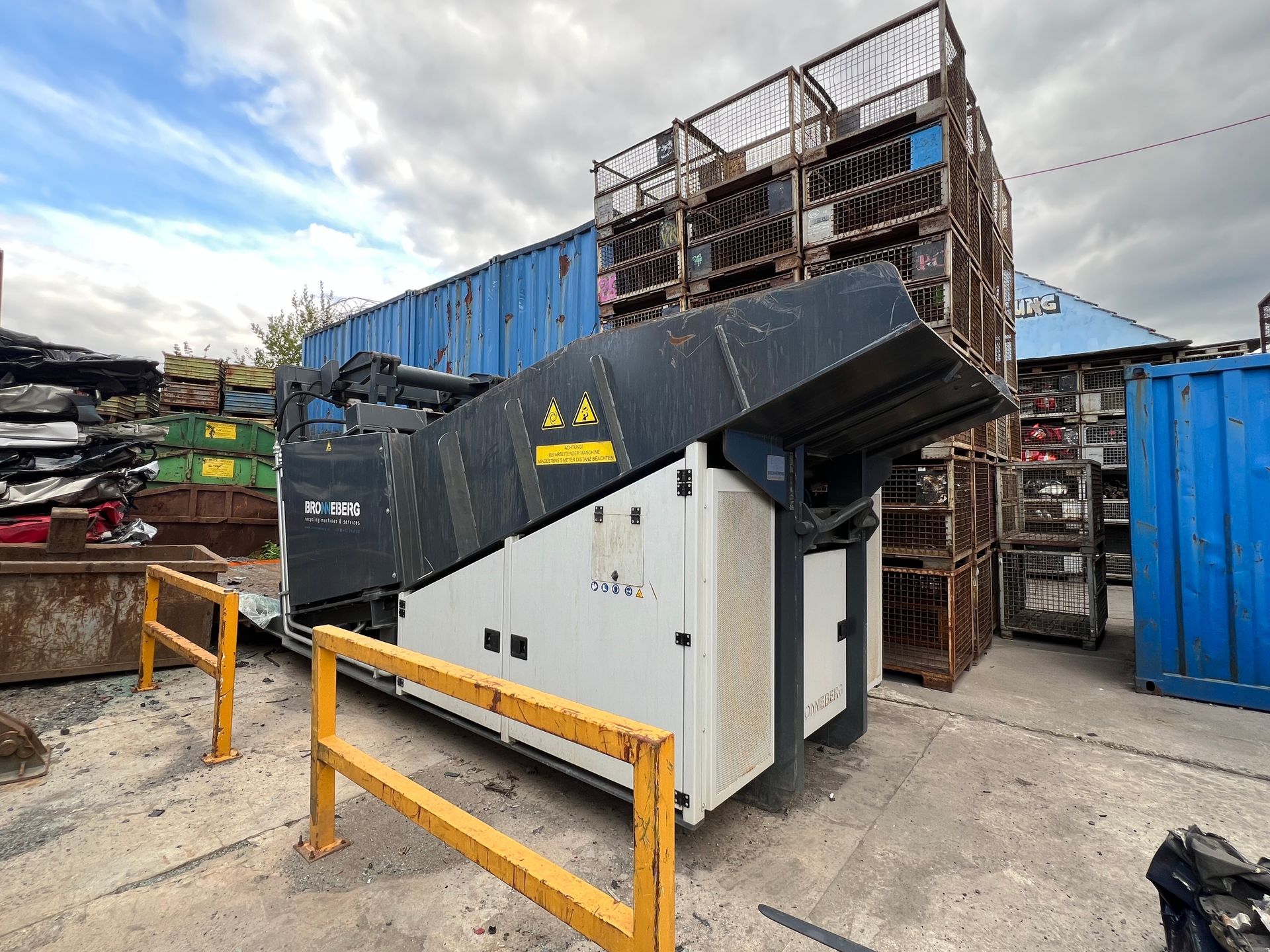 Ein Industriegenerator steht neben einem gestapelten Metallgerüst und einem blauen Container in einem Hof.