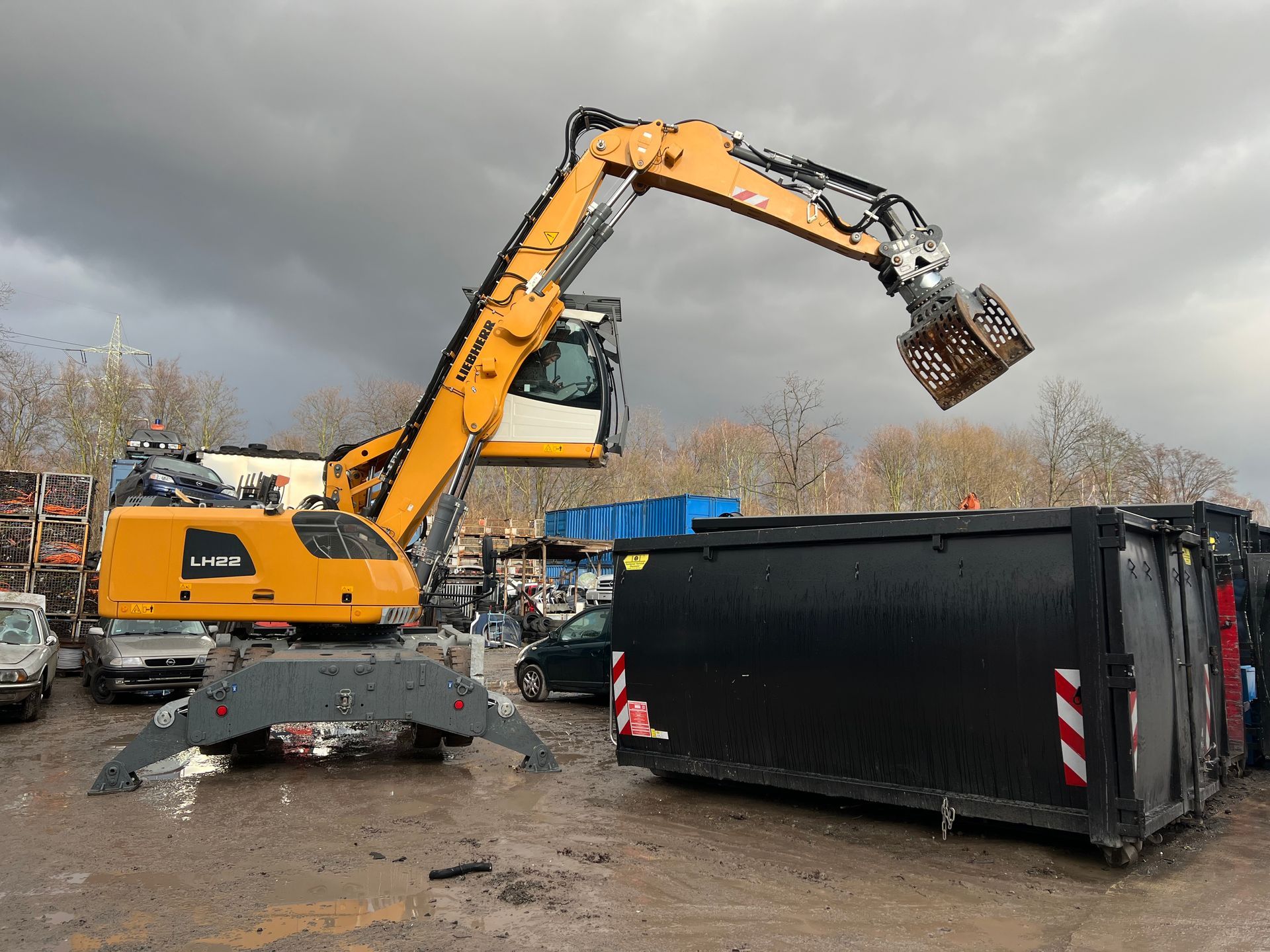 Ein gelber Bagger hebt einen schwarzen Müllcontainer auf einem schlammigen Schrottplatz unter bewölktem Himmel an.