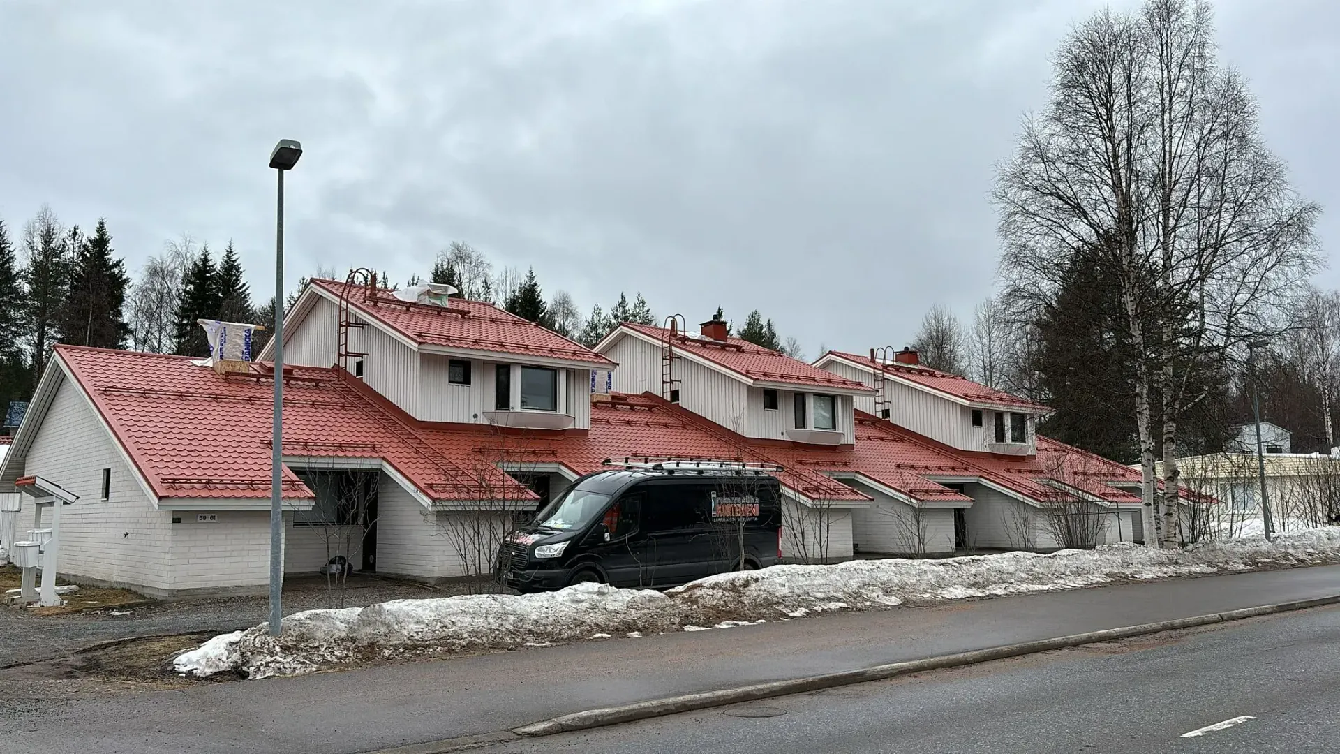 Rivi valkoisia punakattoisia rivitaloja ja musta pakettiauto pysäköitynä edessä pilvisenä päivänä.