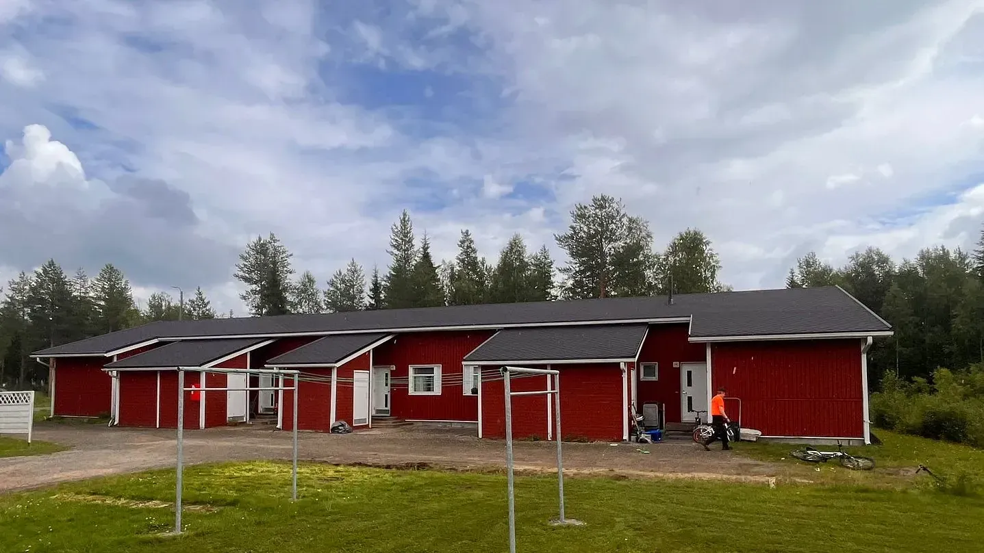 Punaisia rakennuksia pilvisen taivaan alla, puita taustalla, kaksi ihmistä toisen ulkopuolella.
