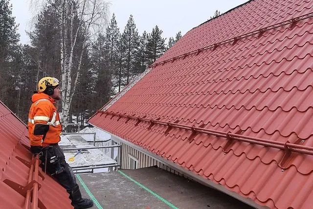 Oranssissa takissa oleva katontekijä tarkastaa punaista kattoa lumisissa ulkotiloissa.