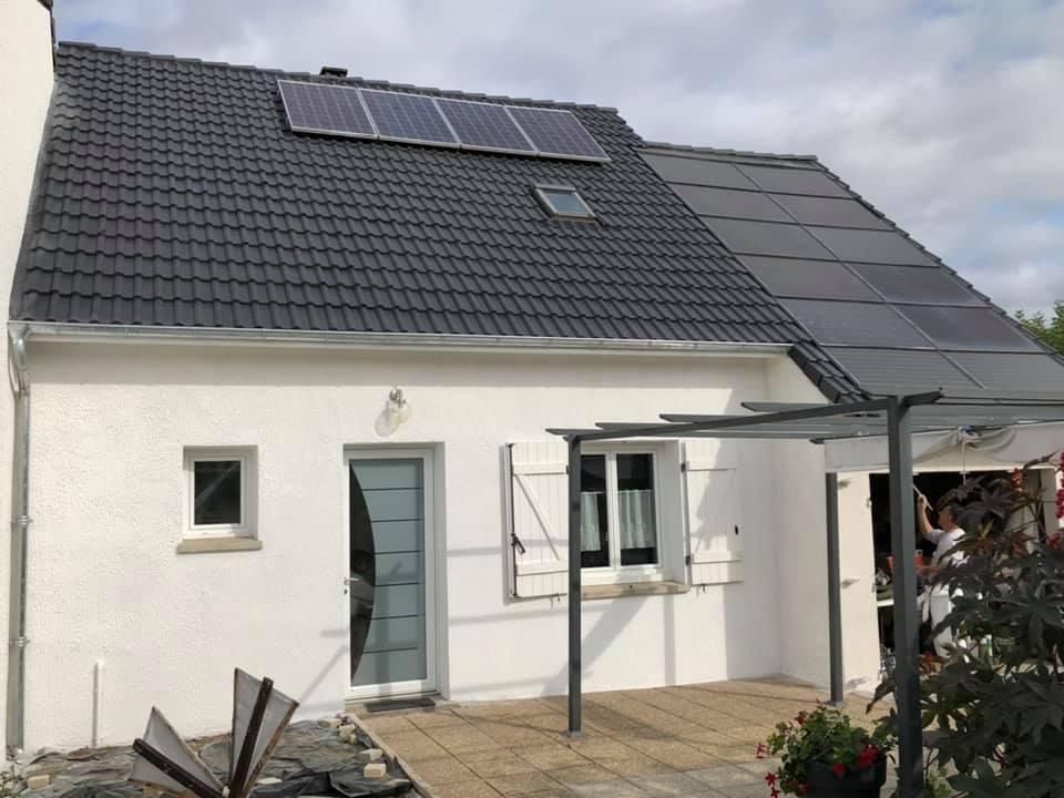 Une maison blanche avec un toit de tuiles sombres, des panneaux solaires et un patio couvert où l'on aperçoit une personne sous la structure.