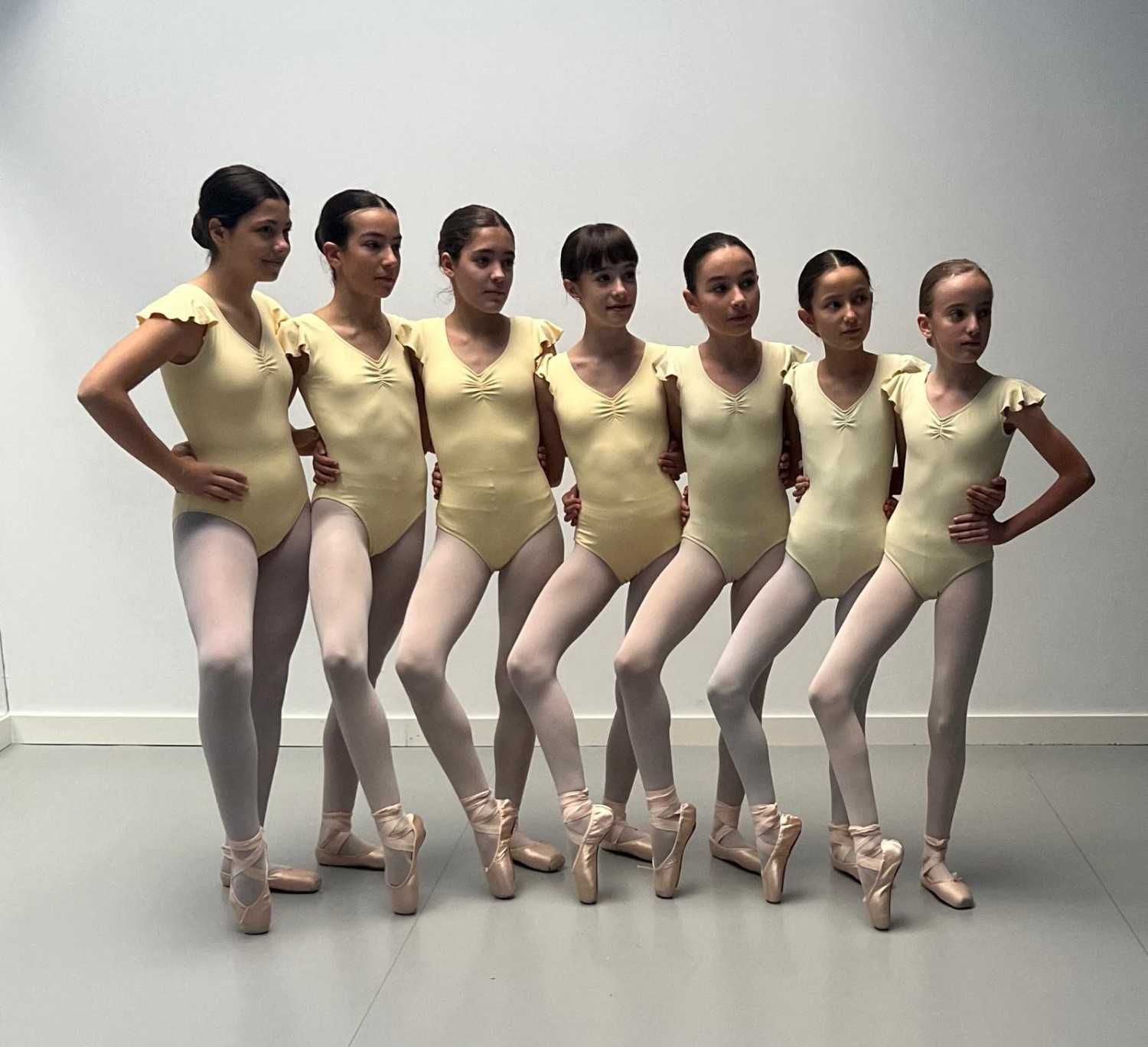 Siete bailarinas de ballet con leotardos amarillo pálido y medias rosas posan de puntillas contra una pared gris.