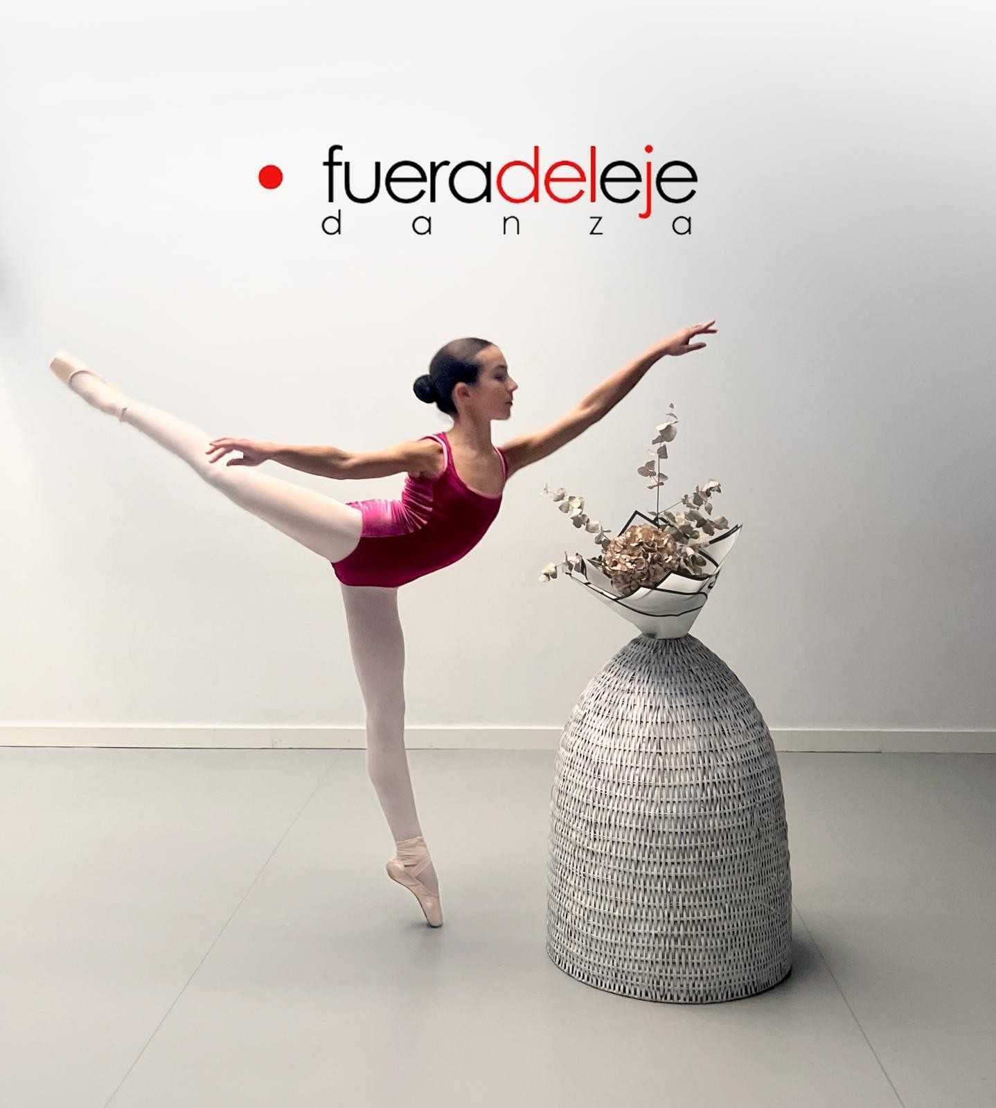 En un estudio, una bailarina de ballet con leotardo rojo se equilibra de puntillas junto a un pedestal gris escultural.