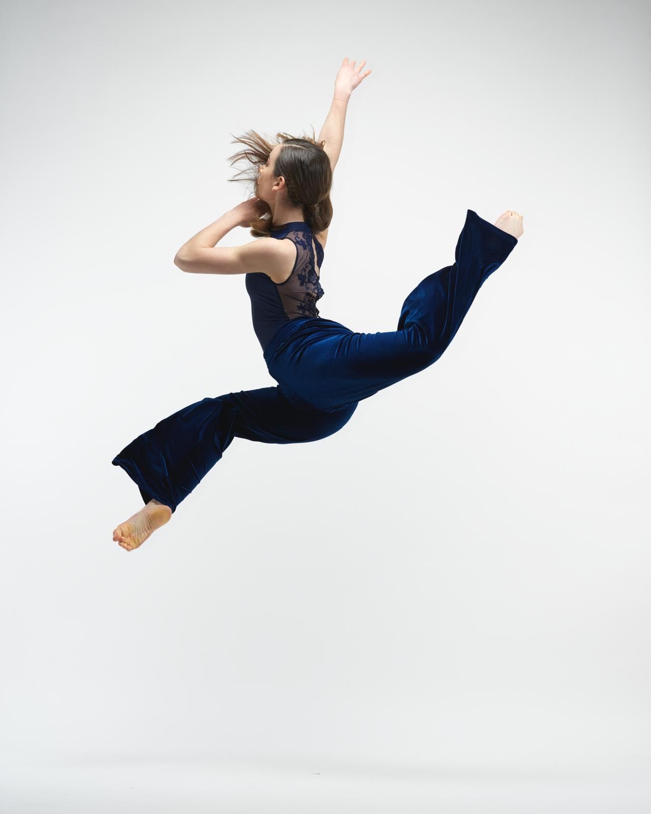 Mujer en el aire realizando un salto dinámico, vistiendo un atuendo azul oscuro sobre un fondo blanco.