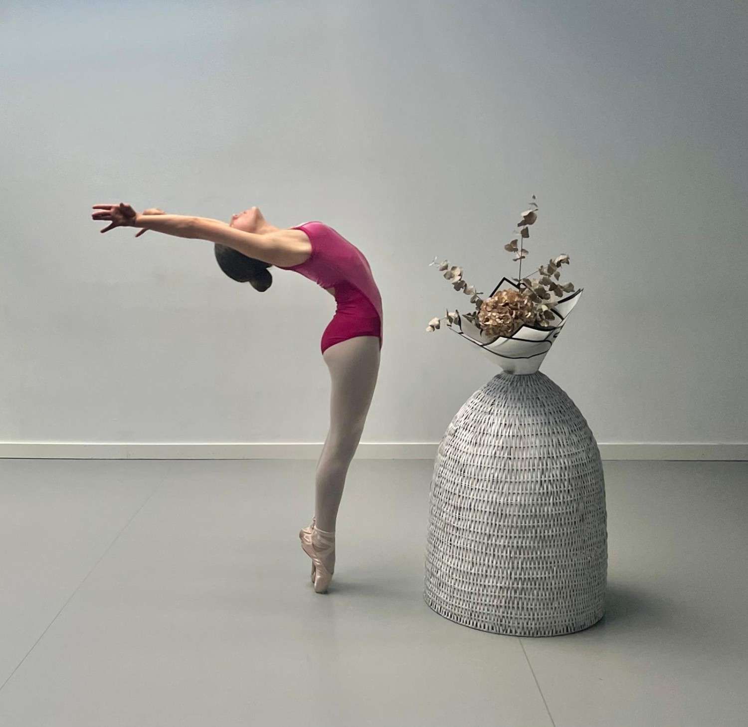 Una bailarina de ballet con leotardo rosa se arquea hacia atrás junto a un gran jarrón gris lleno de flores secas.