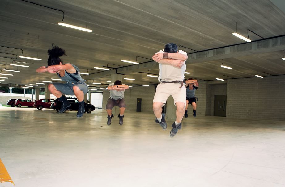 Bailarines en pleno salto en un estacionamiento, borrosos por el movimiento, vestidos con ropa deportiva informal.