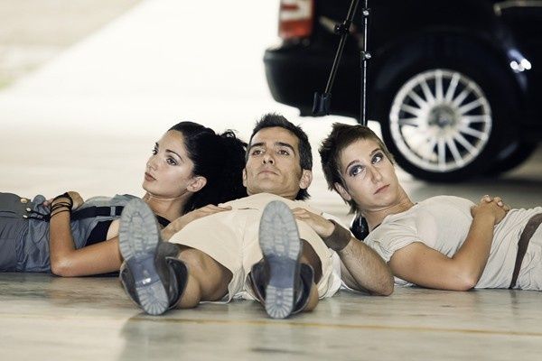 Tres personas tumbadas en el suelo de un garaje, mirando hacia arriba junto a un neumático de coche negro.