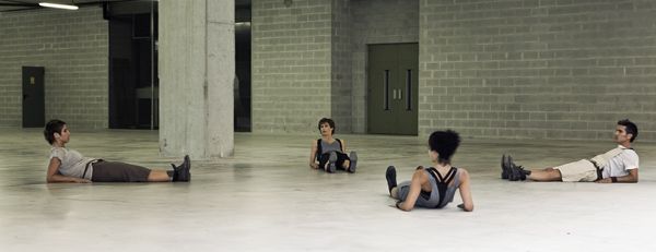 Cuatro personas descansan sobre un suelo de hormigón en una habitación interior austera con columnas y puertas verdes.