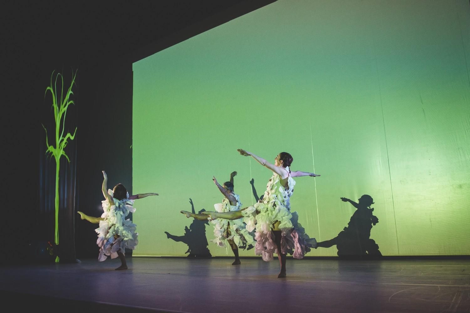 Bailarines con trajes florales actúan en un escenario con un fondo verde brillante y un árbol como elemento decorativo.