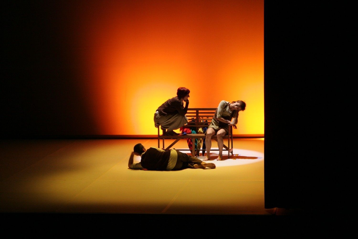 Tres artistas en un escenario iluminado, dos sentados y uno tumbado, con un telón de fondo naranja.