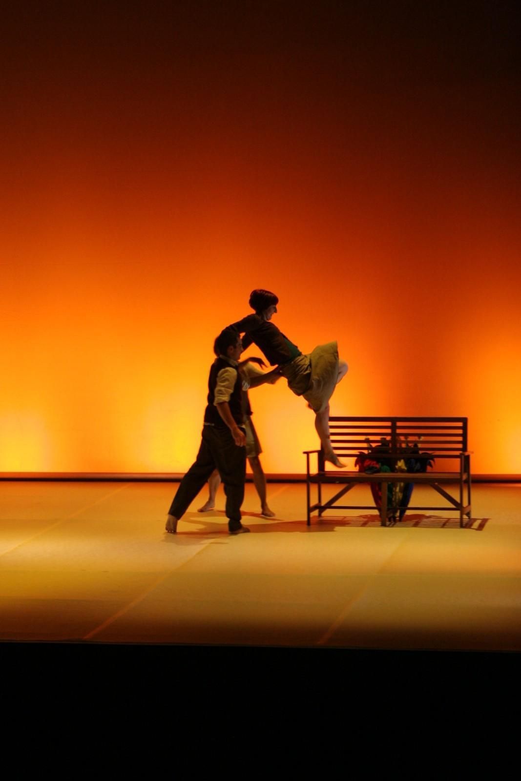 Dos bailarines, silueteados, cerca de un banco en un escenario iluminado con luz naranja.