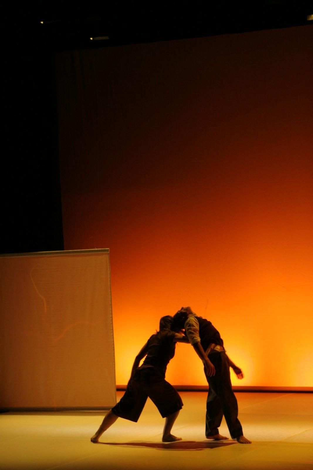 Dos bailarines se inclinan en una pose de pareja sobre un escenario con un fondo naranja brillante.