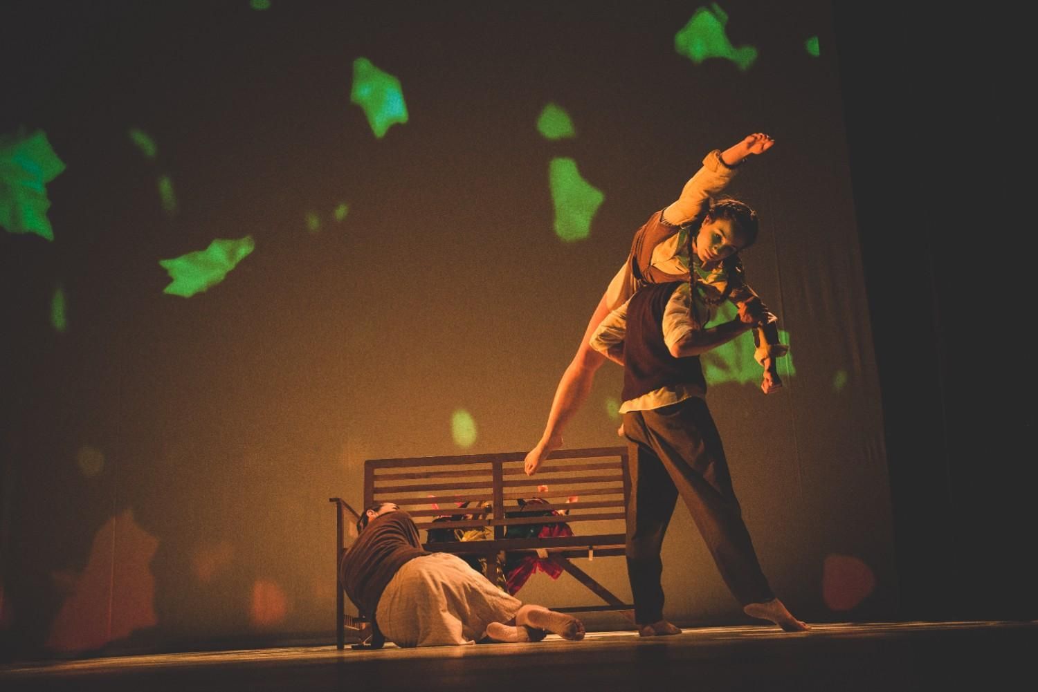 Escena teatral con una persona recostada sobre otra de pie junto a un banco bajo una iluminación cálida y dramática.