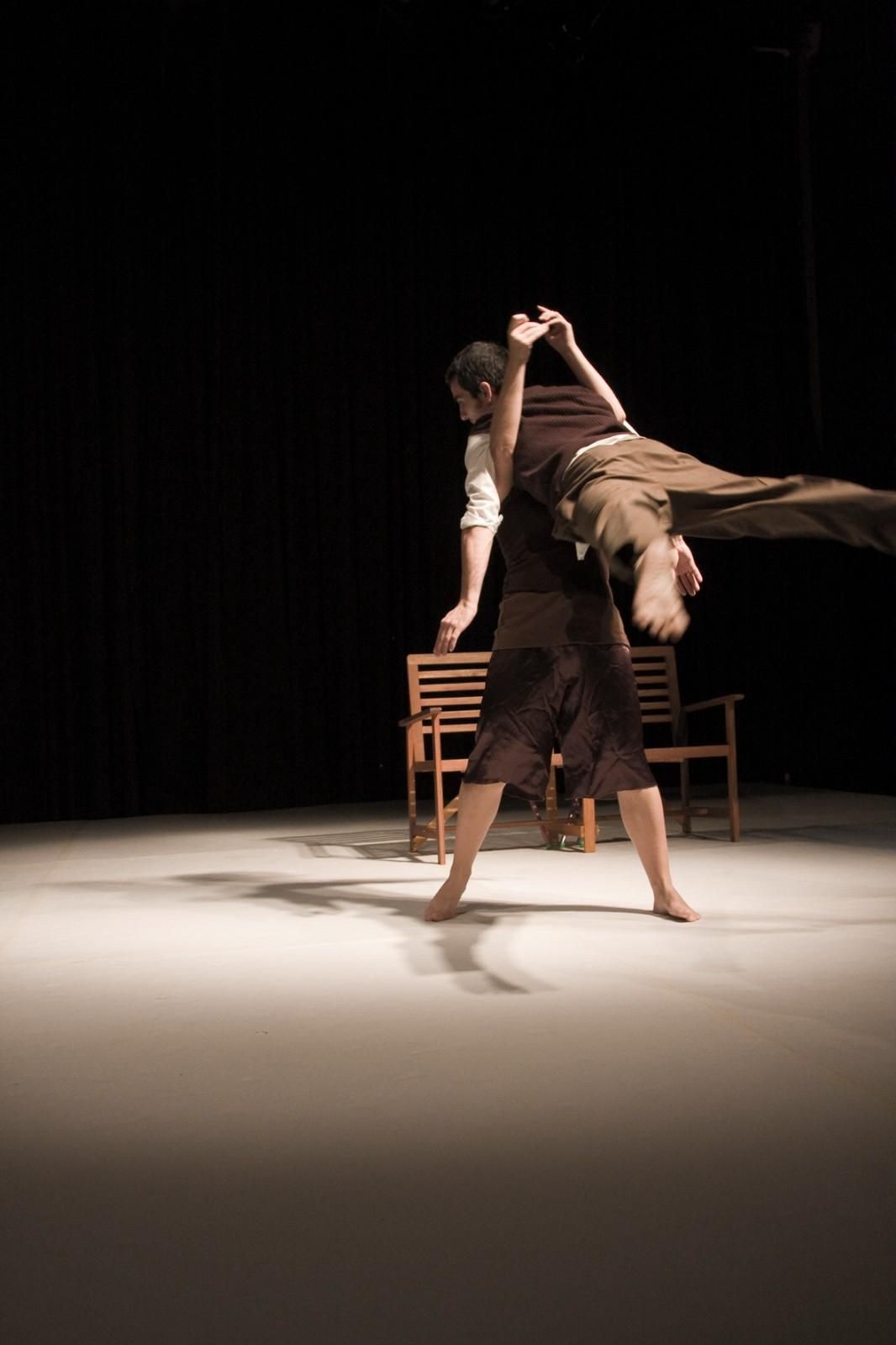 Dos bailarines en una actuación en un escenario con poca luz, uno levantando al otro por encima de una silla y una mesa.