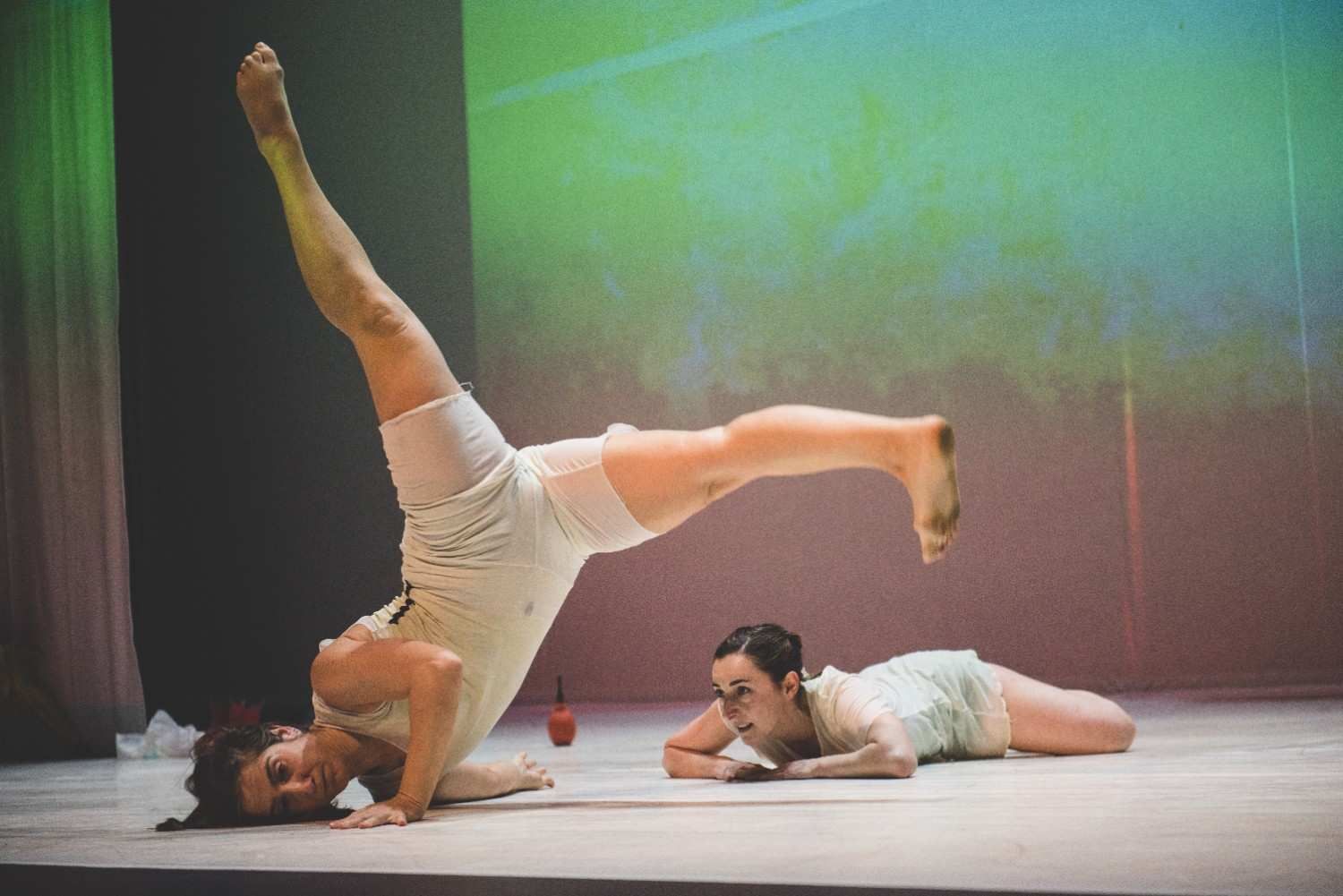 Dos artistas vestidos de blanco en el escenario, uno de ellos haciendo equilibrio boca abajo mientras el otro yace cerca.