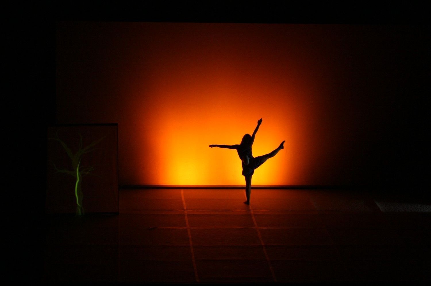 Silueta de una bailarina en equilibrio, posando frente a un foco naranja brillante en un escenario oscuro.