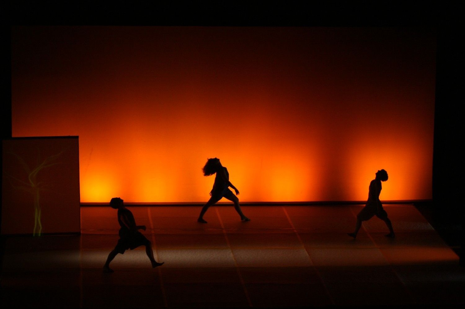 Tres bailarines en silueta sobre un escenario iluminado por un telón de fondo naranja brillante.