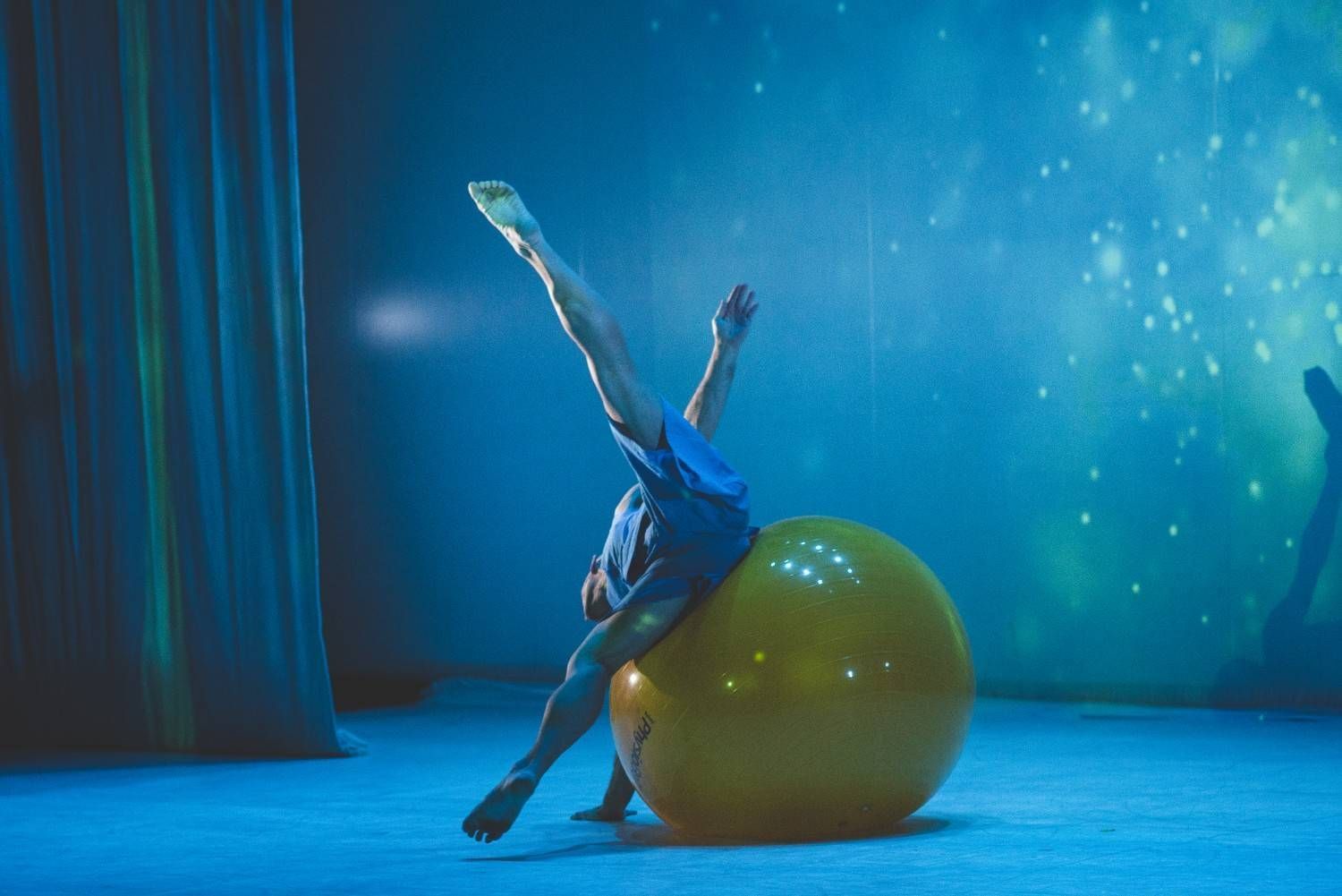Una bailarina vestida de azul posa sobre un brazo junto a una gran pelota verde en un escenario con poca luz.